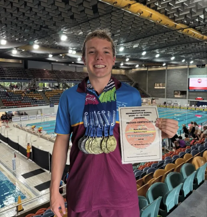 A young swimmer at a championship pool holding up a certificate, a row of medals around his neck