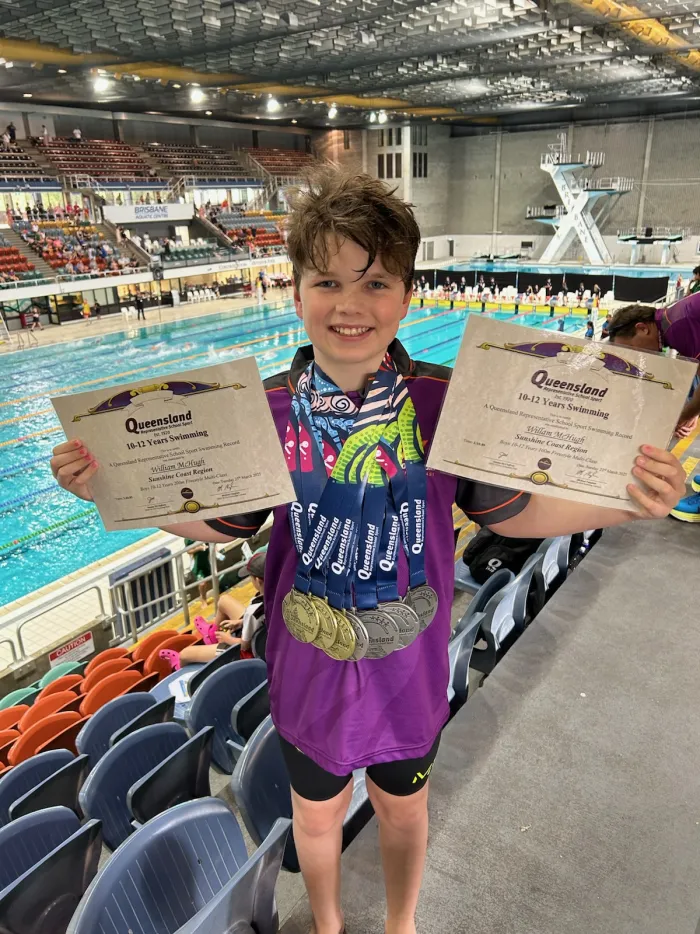 A junior swimmer holding two certificates, medals around his neck, in the stands above an indoor pool